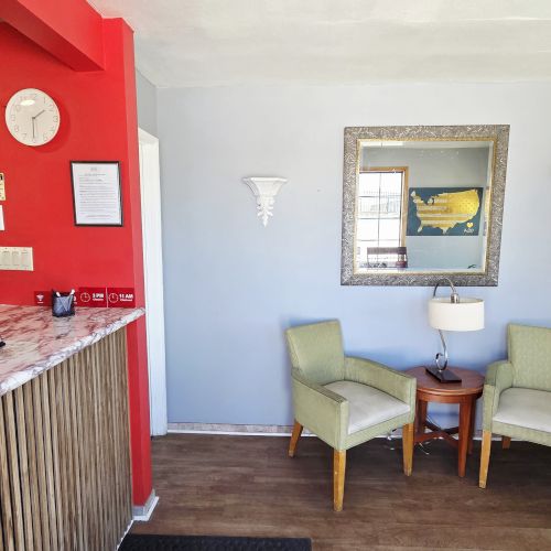 The image shows a small reception area with a red wall, chairs, a table, a mirror, and a coffee maker on a desk.