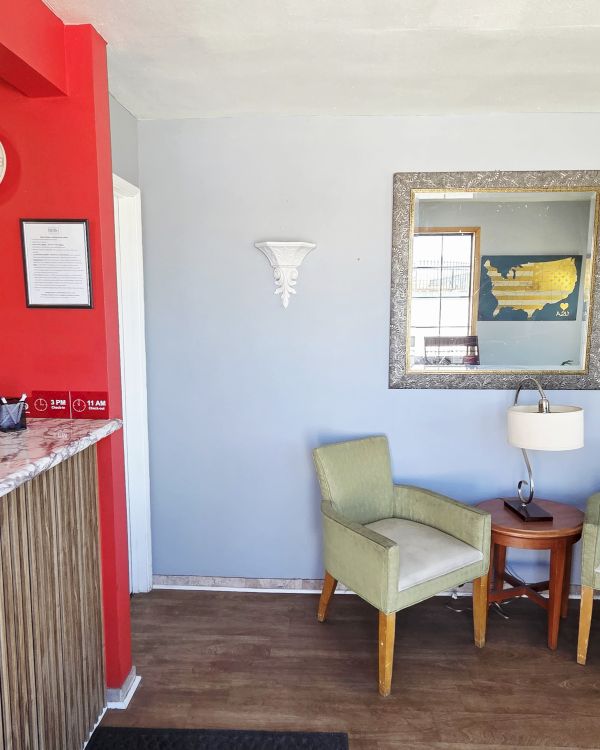 The image shows a small reception area with a red wall, chairs, a table, a mirror, and a coffee maker on a desk.