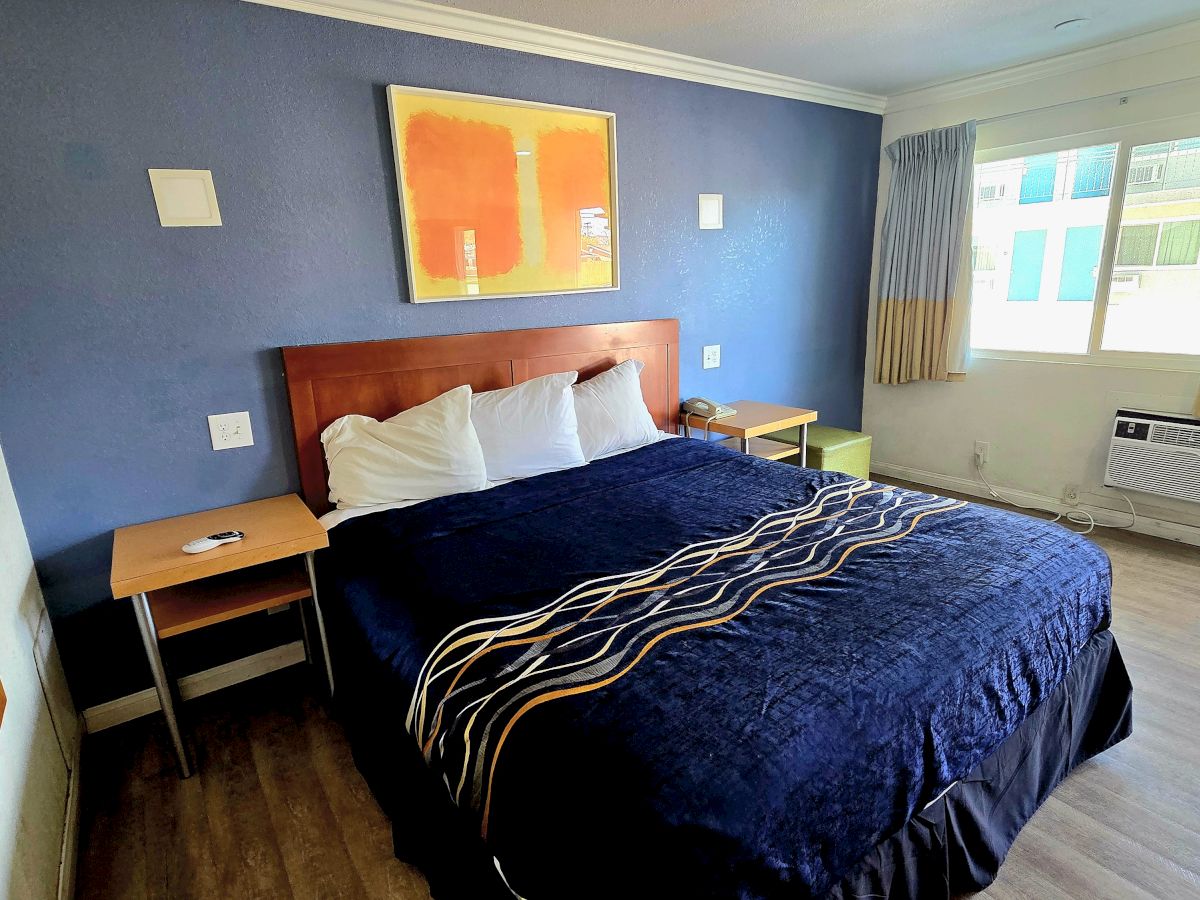 A modern hotel room features a double bed with a dark blanket, blue wall, abstract art, bedside tables, window, and air conditioner.
