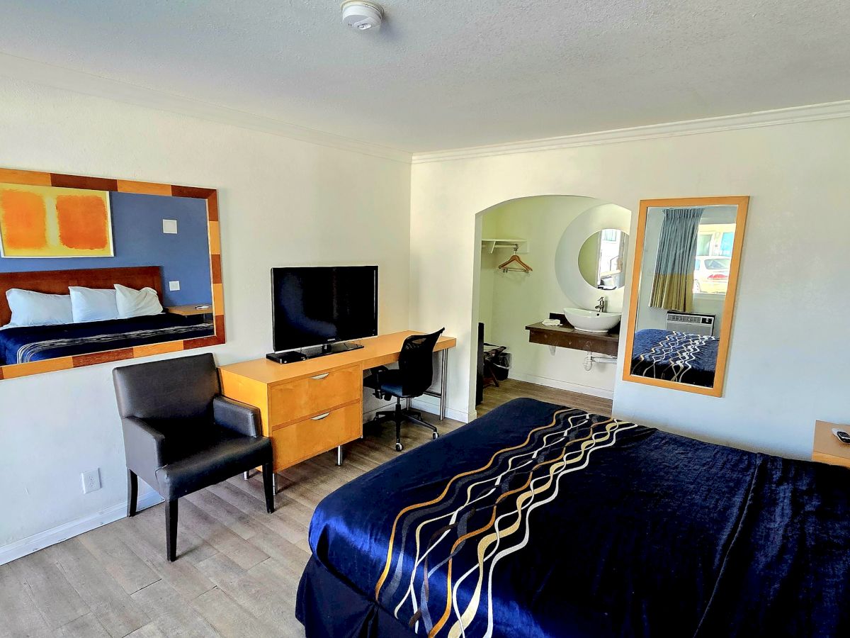 The image shows a hotel room with a bed, desk, chair, TV, and large mirror, styled in a modern decor with blue and wooden accents.