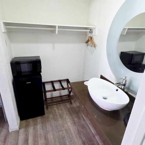 The image shows a room corner with a sink, a mirror, a microwave, and shelves. Floor is wooden and space appears minimalistic.