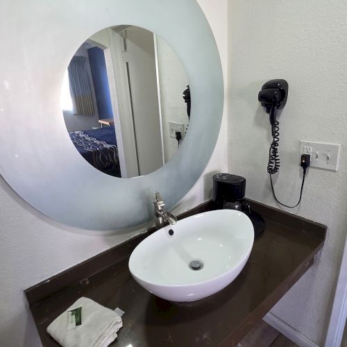 A bathroom sink with a round mirror, hairdryer, towel, and soap on a dark countertop, with a wooden floor.