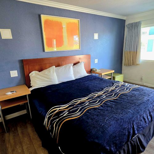 A hotel room features a bed with blue bedding, a window, a wall-mounted artwork, nightstands, and an air conditioner unit by the window.