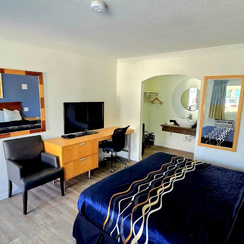 A hotel room with a bed, TV, desk, chair, and large mirror. There's a blue accent wall and an open archway leading to another area.