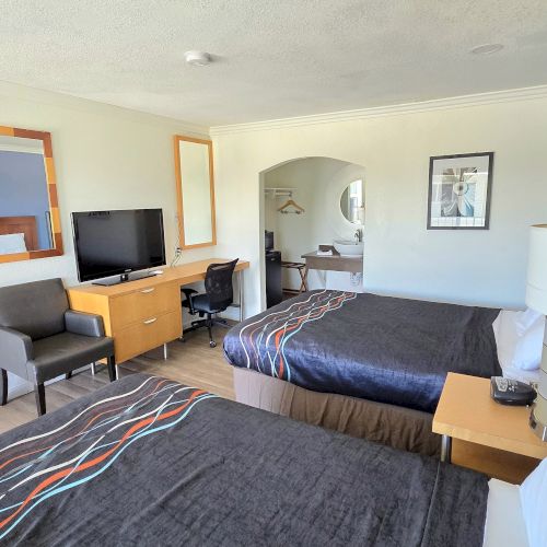 The image shows a hotel room with two beds, a TV, a desk, and a chair. The decor is modern with a large mirror on the wall.