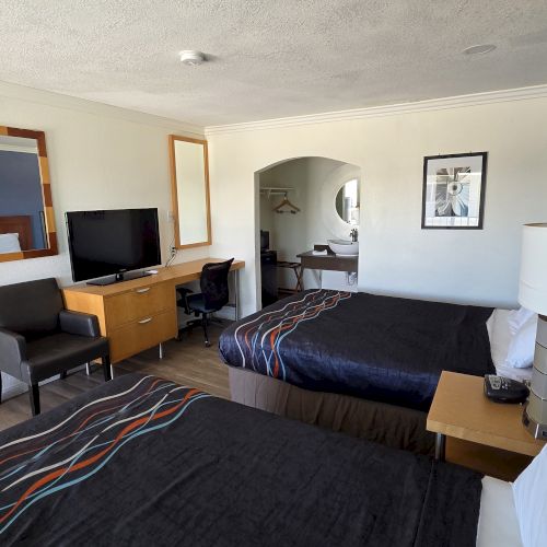 The image shows a hotel room with two beds, a TV, a desk, and a chair. The room has a modern, simple decor with framed artwork.