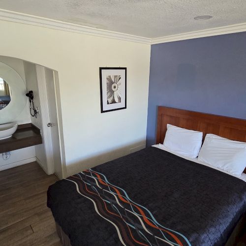 A hotel room with a bed, bedside table, lamp, and artwork; a sink and mirror are visible in an adjacent alcove.