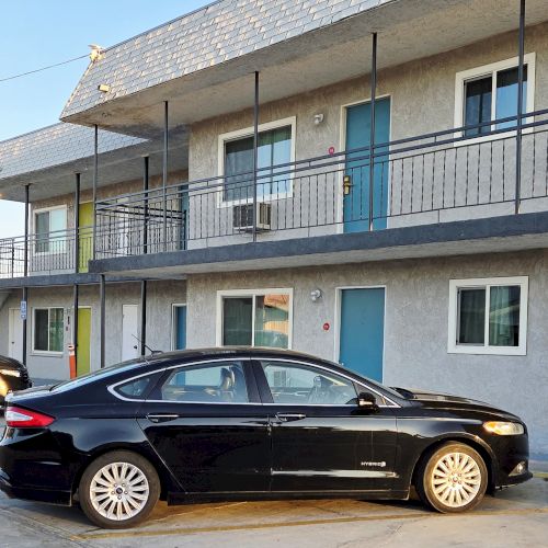 The image shows a small apartment building or motel with two parked cars in front, featuring a stairway and outdoor corridors.