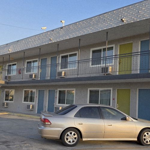The image shows a two-story motel building with a row of doors and windows, featuring parked cars in the foreground.