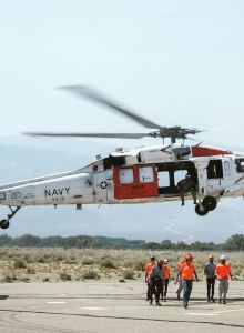 A helicopter labeled "Navy" hovers above the ground as several people wearing orange vests walk nearby on a tarmac in an arid area.