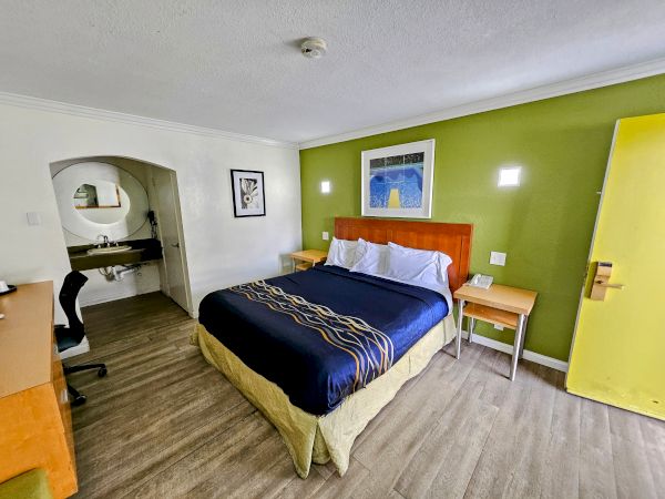 A hotel room with a bed, desk, and artwork on the wall. There is a sink area to the left with an arched opening.