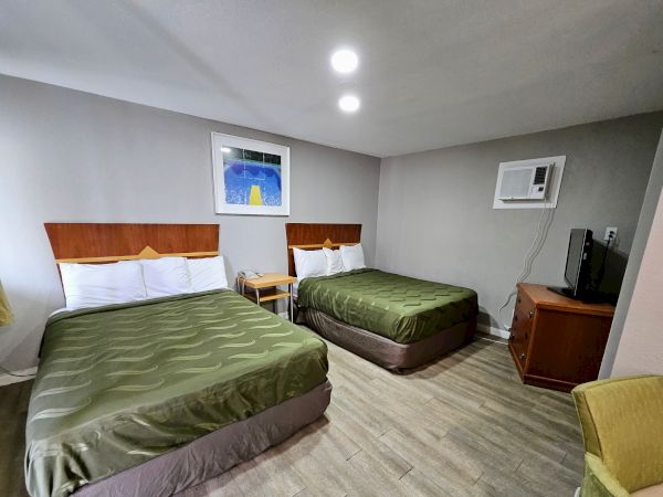 The image shows a hotel room with two double beds, a nightstand, wall art, an air conditioner, a TV on a wooden cabinet, and a green armchair.