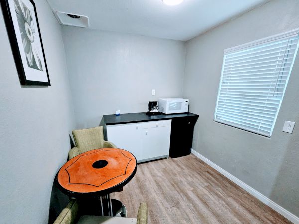 A small room with a table and two chairs, a kitchenette with a microwave and coffee maker, and a window with closed blinds.