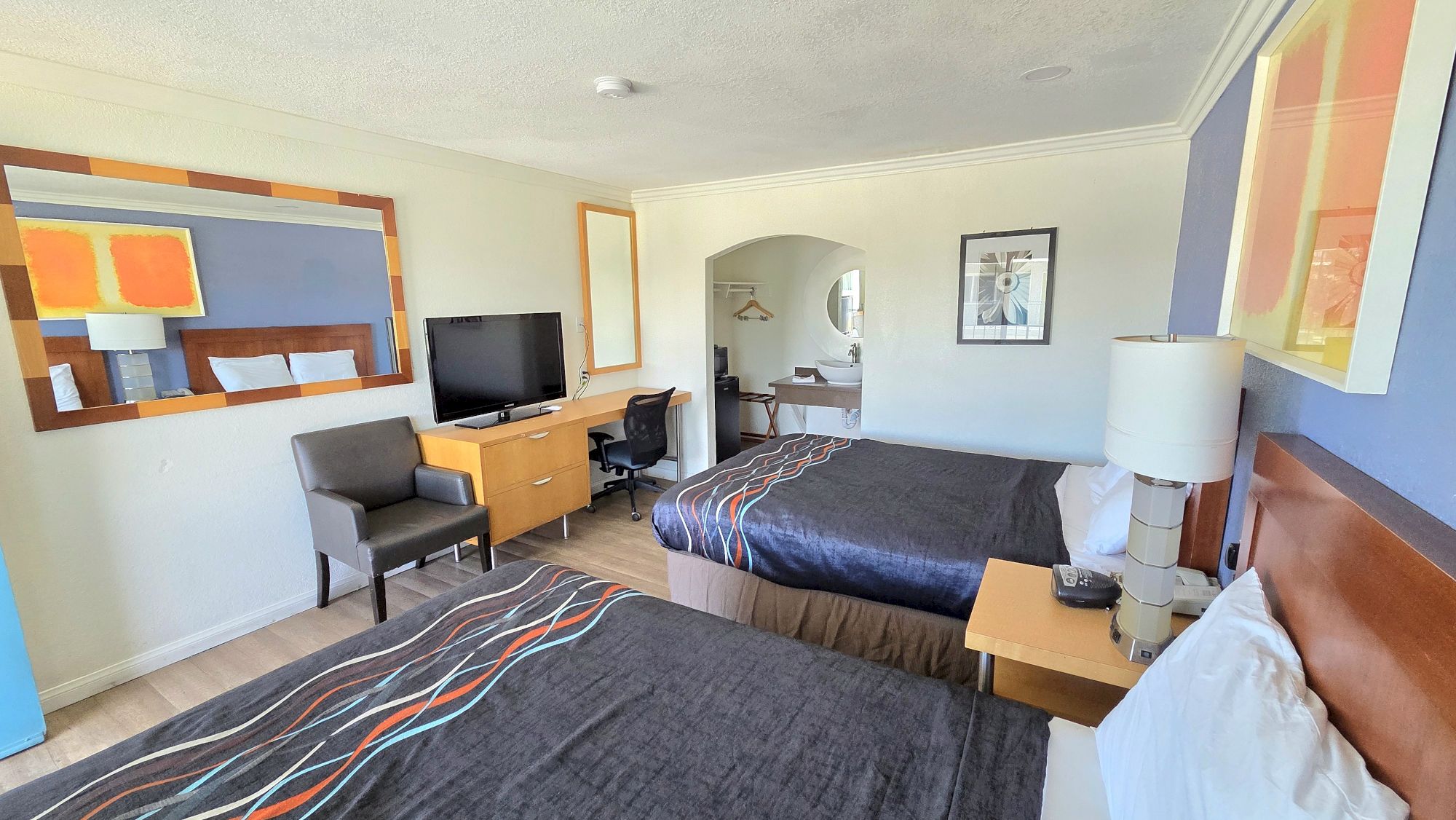 The image shows a hotel room with two double beds, a TV, a desk, a chair, and modern decor.