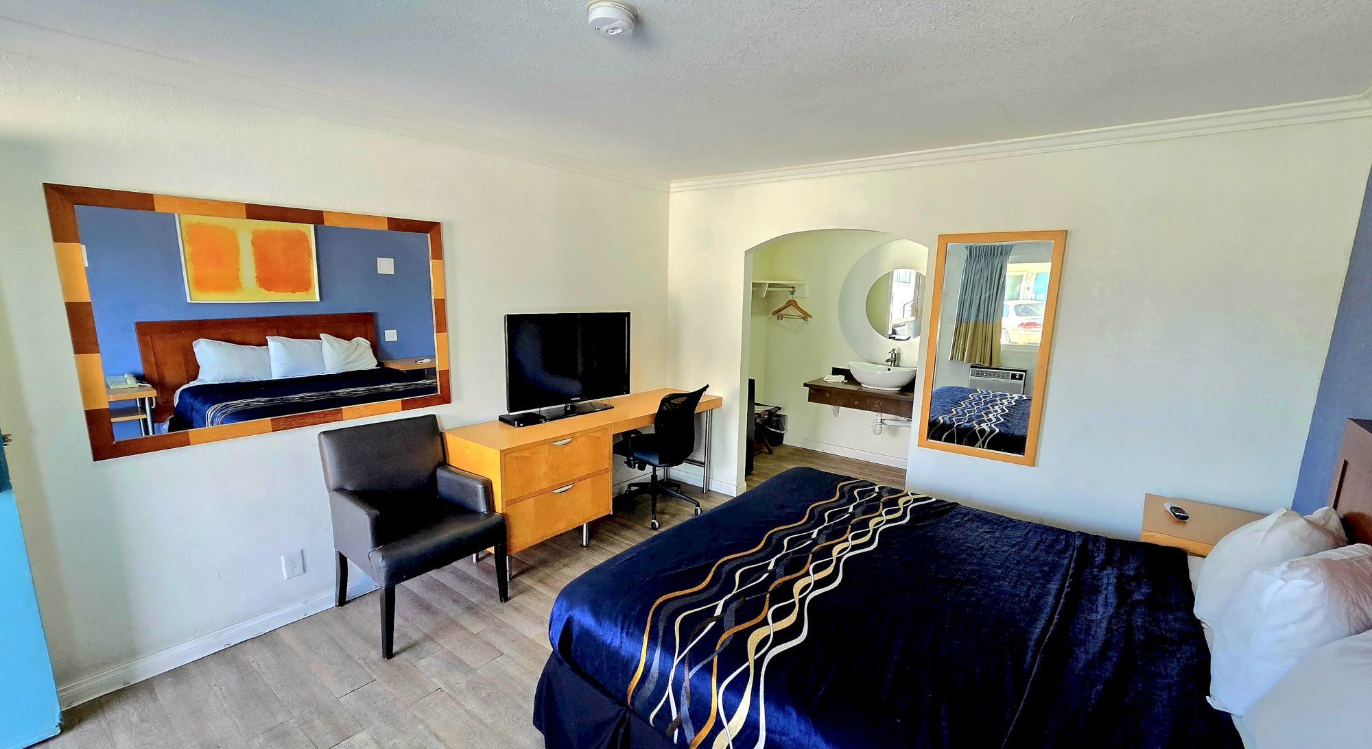 The image shows a hotel room with a bed, desk, chair, TV, wall art, and a mirror. There's a separate area visible through an archway.